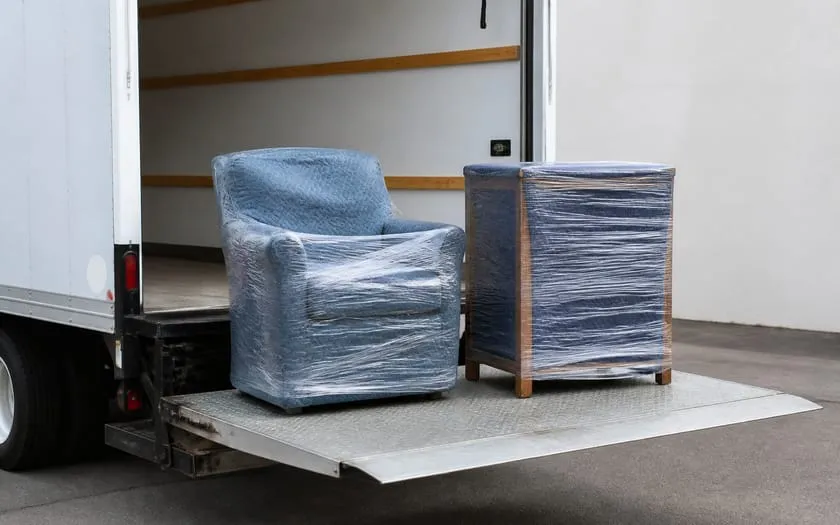In Umzugsdecken und Stretchfolie verpackter Sessel und Holzschrank auf der Ladebordwand eines Lkw